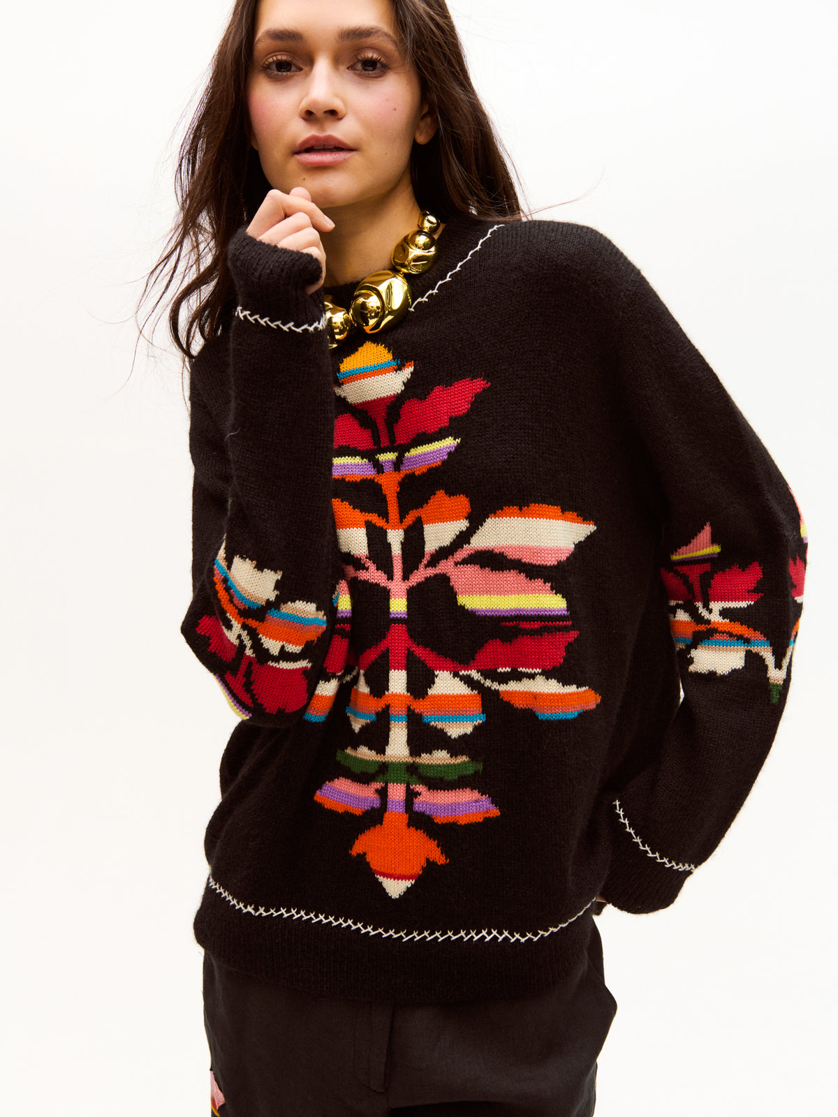 Model wearing a black sweater with a colorful abstract floral pattern in shades of red, orange, pink, and blue, styled with a bold gold statement necklace. The look blends modern artistry with timeless craftsmanship.