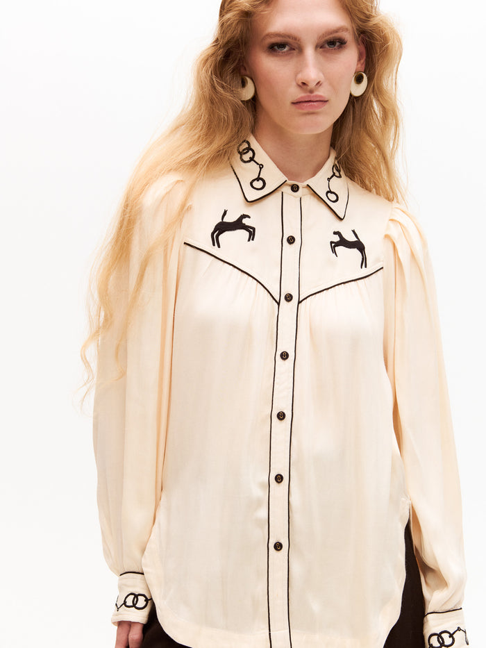 Model wearing a cream long-sleeve blouse with black embroidery on the collar and chest. The shirt features horse motifs and black piping details along the seams and buttons.