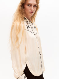 Model in a cream blouse looking over her shoulder. The front and side details, including black horse embroidery, piped seams, and cuff detailing, are partially visible.
