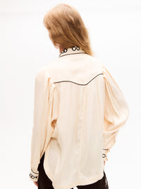 Rear view of the cream blouse displaying a soft drape, pleated back detail, and black piping outlining the yoke. Embroidered chain detail is visible on the collar.