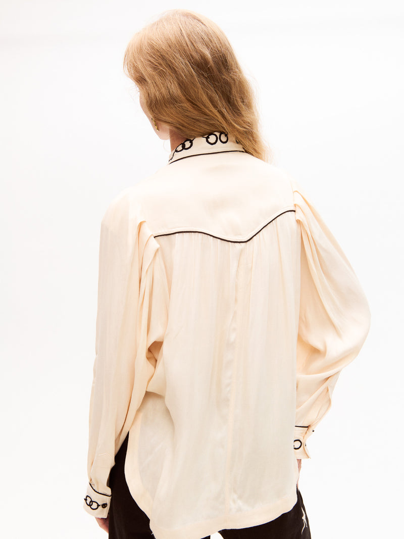 Rear view of the cream blouse displaying a soft drape, pleated back detail, and black piping outlining the yoke. Embroidered chain detail is visible on the collar.