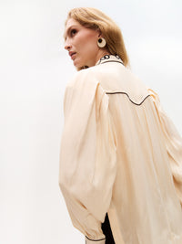 Side angle showing the voluminous sleeve and shoulder pleats of the cream blouse, with embroidered black chain motifs on the collar. The model gazes to the side, highlighting the earring.