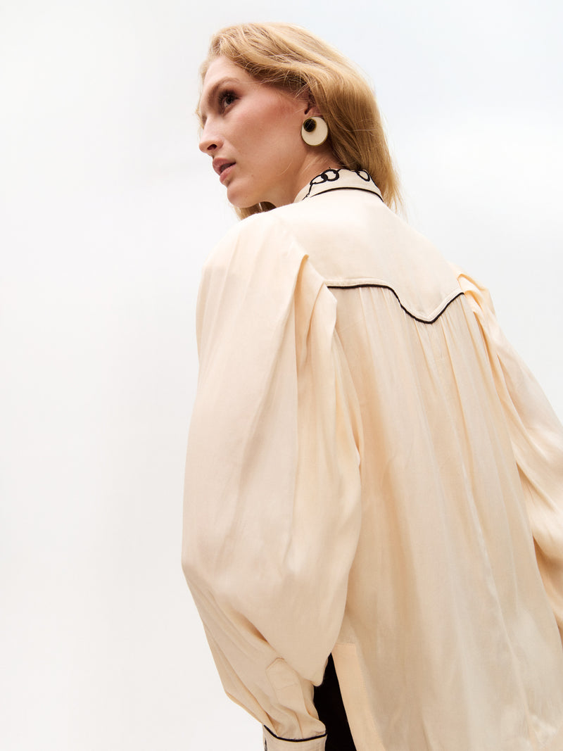 Side angle showing the voluminous sleeve and shoulder pleats of the cream blouse, with embroidered black chain motifs on the collar. The model gazes to the side, highlighting the earring.