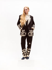 A model stands wearing a striking black suit with contrasting cream embroidery, including a structured blazer and tailored trousers. The ensemble is paired with a cream western-style blouse featuring black piping and embroidery, completed with blue heeled shoes.