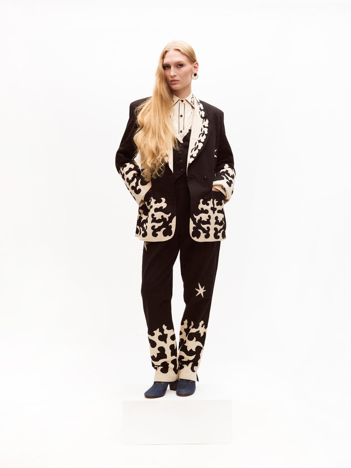 A model stands wearing a striking black suit with contrasting cream embroidery, including a structured blazer and tailored trousers. The ensemble is paired with a cream western-style blouse featuring black piping and embroidery, completed with blue heeled shoes.