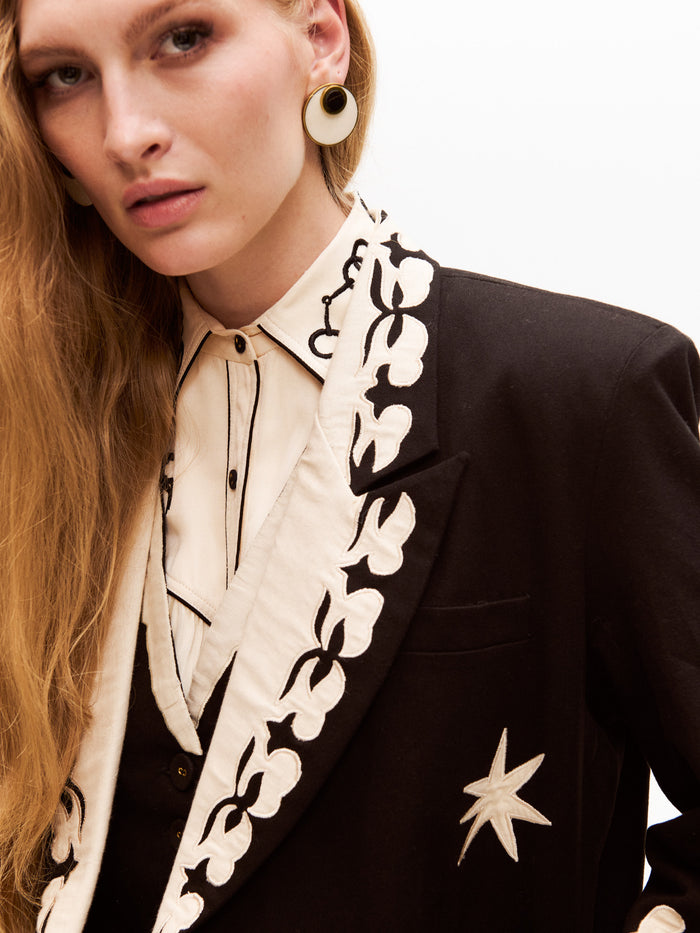 Close-up view of the model’s upper torso showing the detailed embroidery on the cream shirt collar and the lapel of the blazer. The blazer features star embroidery and a delicate floral appliqué along the lapels.