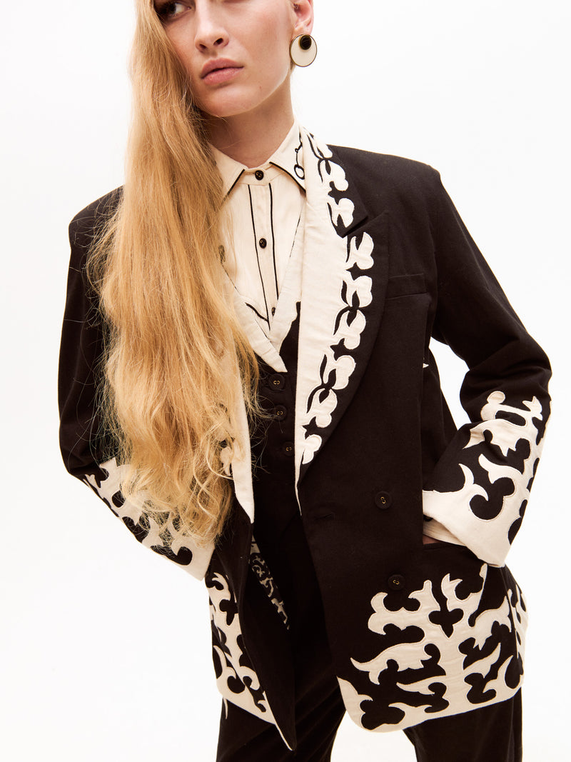 The model poses with one hand in her pocket, showcasing the elaborate embroidery on the sleeves and hem of the blazer, along with matching cream details on the pants.