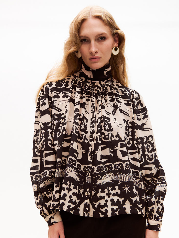 Close-up of the same model wearing the printed blouse, showcasing the pleated neckline and whimsical pattern in detail. Her expression is poised, and the high-neck design continues under the chin for a structured, elegant look.