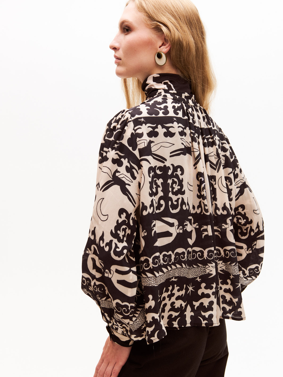 Side view of the model revealing the loose fit and voluminous back of the patterned blouse. The whimsical print wraps around the blouse, emphasizing the artistic, hand-drawn look of the dragons, moons, and horses.