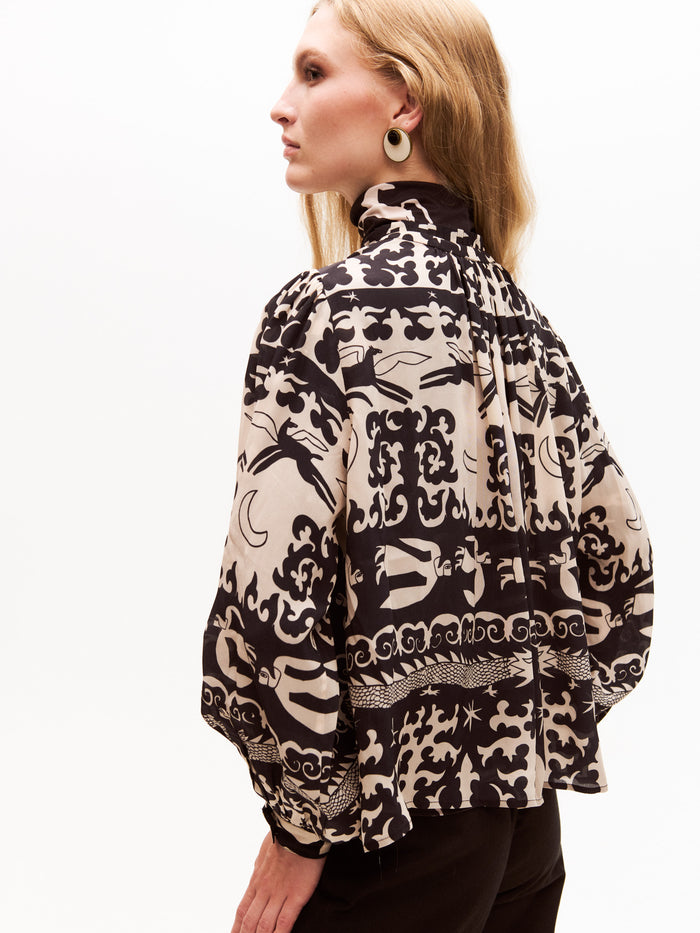Side view of the model revealing the loose fit and voluminous back of the patterned blouse. The whimsical print wraps around the blouse, emphasizing the artistic, hand-drawn look of the dragons, moons, and horses.