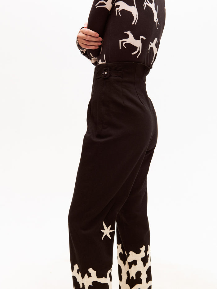 Back view of a woman wearing high-waisted black trousers with cream appliqué at the bottom, paired with a black long-sleeve top patterned with cream galloping horses. Her arms are crossed over her chest.
