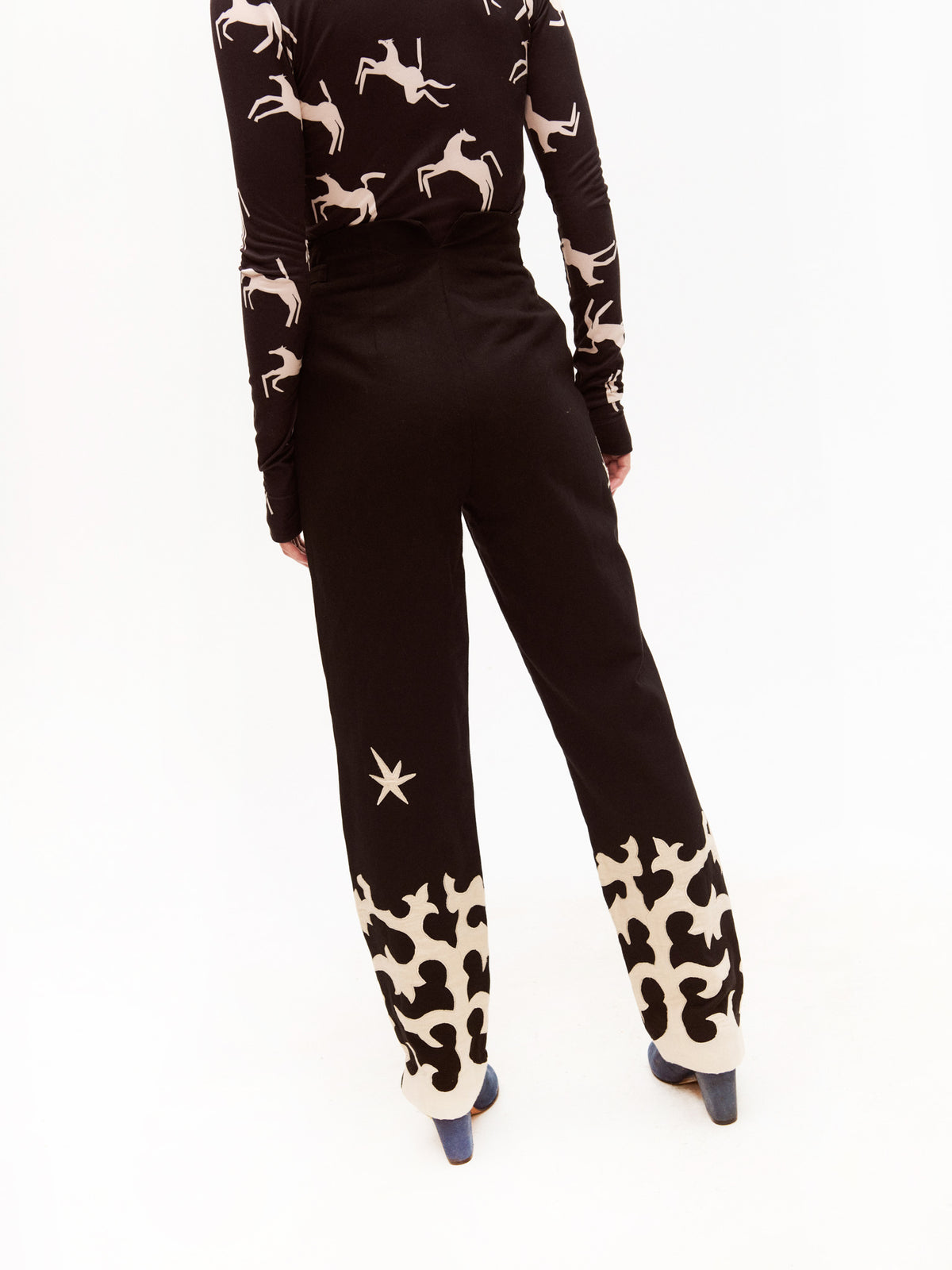 Full back view of a woman wearing the same black high-waisted trousers with cream appliqué and star embroidery, styled with matching blue heeled shoes and a long-sleeve printed top featuring cream horse silhouettes on black.