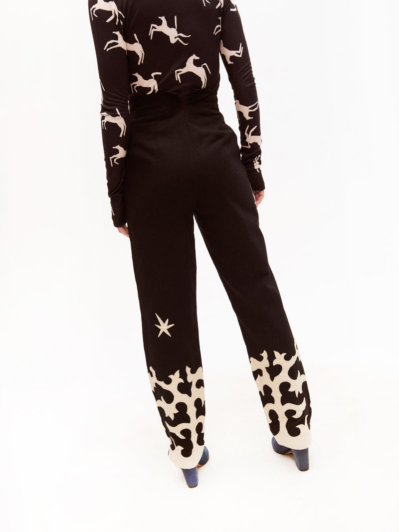 Full back view of a woman wearing the same black high-waisted trousers with cream appliqué and star embroidery, styled with matching blue heeled shoes and a long-sleeve printed top featuring cream horse silhouettes on black.