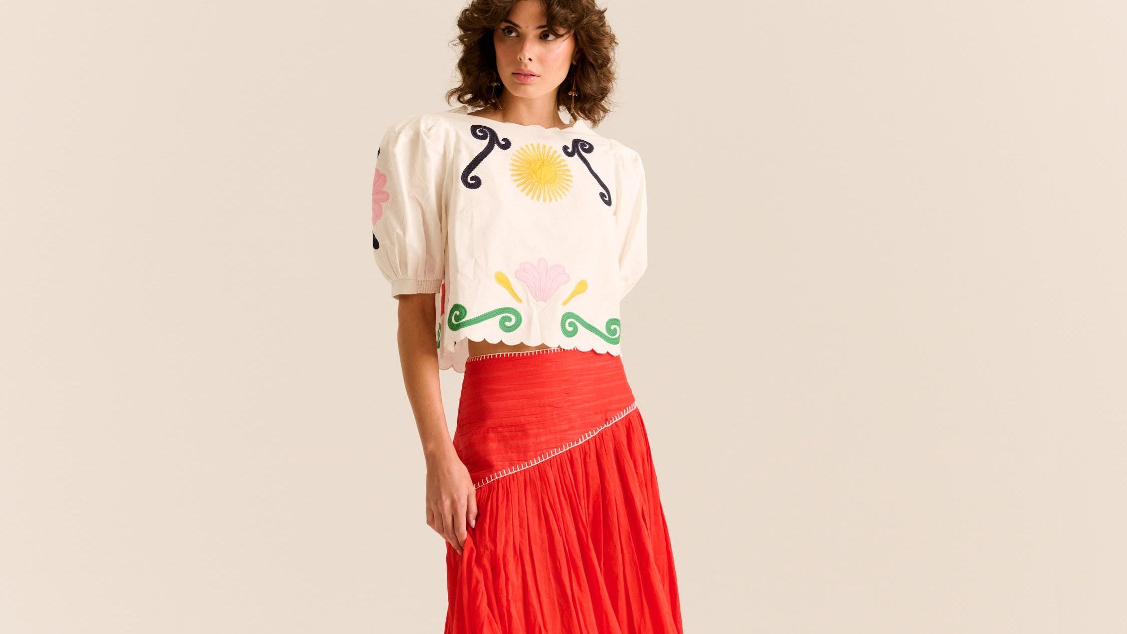 Woman wearing a white blouse with colorful designs and a red skirt on a beige background