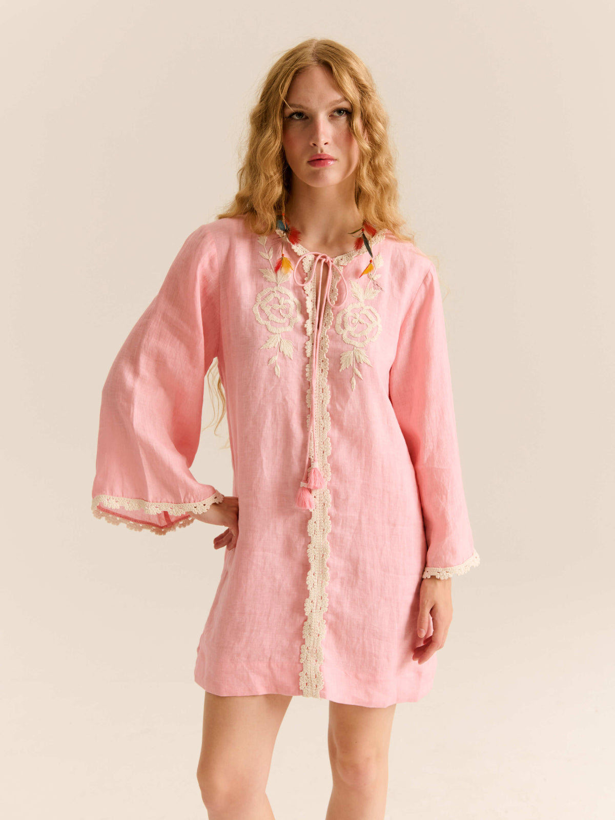 Model wearing a pink long-sleeve mini dress with lace trim, floral embroidery on the chest, and a tassel tie neckline.