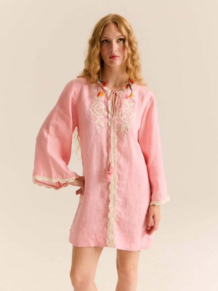Model wearing a pink long-sleeve mini dress with lace trim, floral embroidery on the chest, and a tassel tie neckline.