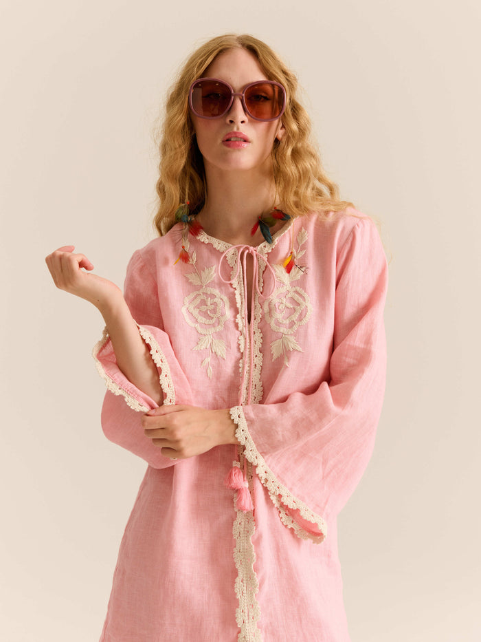 Close-up of the pink embroidered mini dress featuring lace trim, floral embroidery at the neckline, tassel ties, and the model wearing oversized sunglasses and colorful feather earrings.