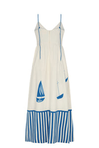 White maxi dress with thin adjustable blue straps, a flowing skirt, and blue sailboat motifs. Features a striped blue-and-white hem and an open V-back design.