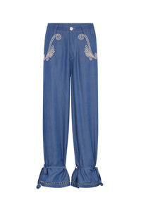 Relaxed-fit denim pants featuring embroidered detailing at the front pockets, a button closure, and adjustable ankle ties with tassel accents. Finished with decorative trim at the cuffs for a handcrafted touch.