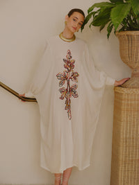 Model wearing a flowing ivory kaftan dress with a colorful embroidered motif down the front, styled with a gold collar necklace beside a staircase and woven plant stand.