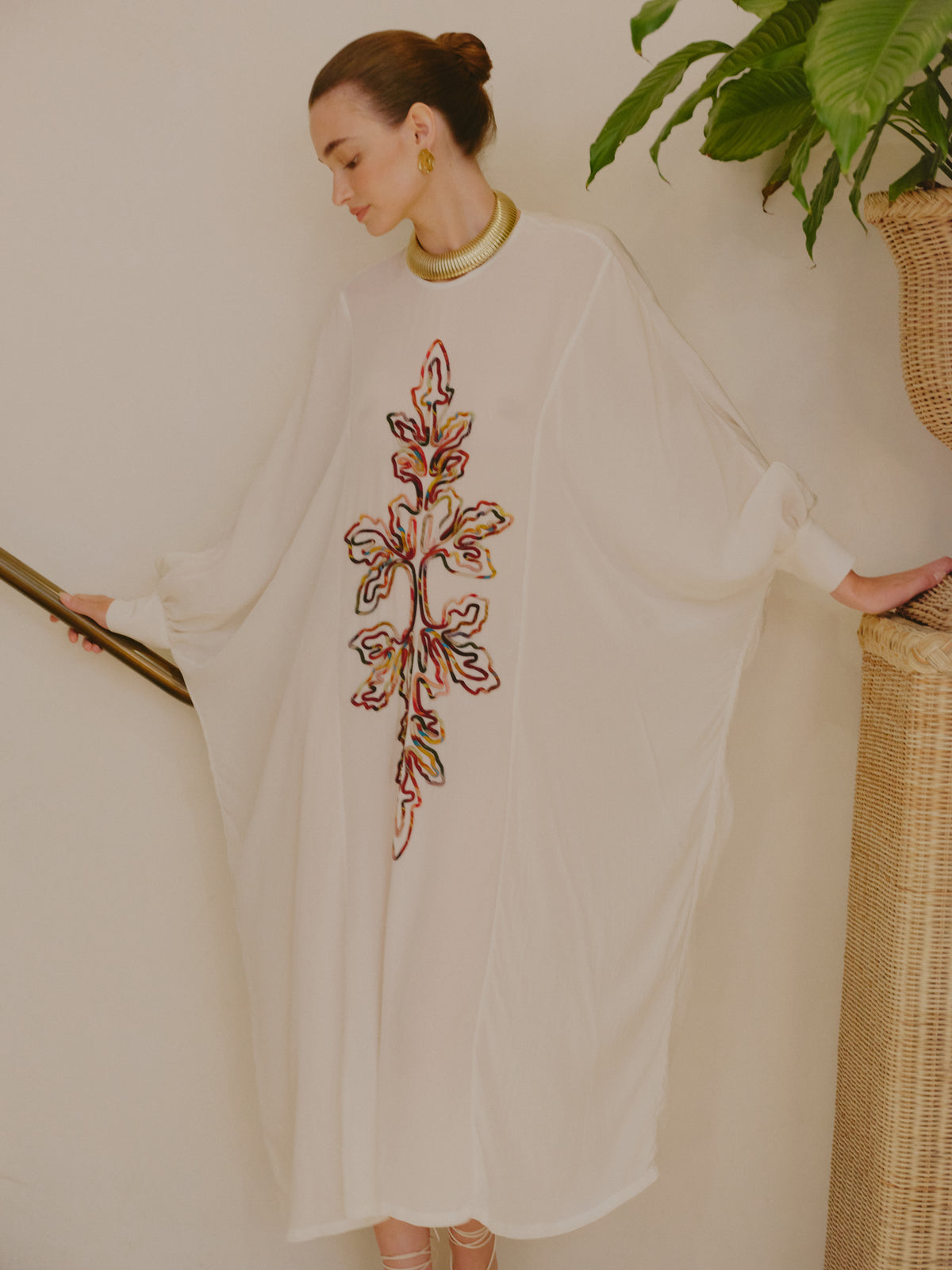 Side view of a model in an ivory embroidered kaftan dress with wide sleeves, styled with a gold necklace and earrings.
