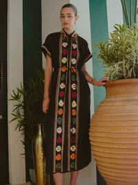 Model wearing a black midi dress with colorful embroidered vertical panels, standing beside a large ceramic planter.