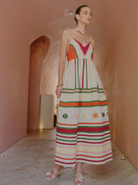 Model wearing a multicolored striped maxi dress with thin straps and embroidered motifs, standing in a curved corridor.