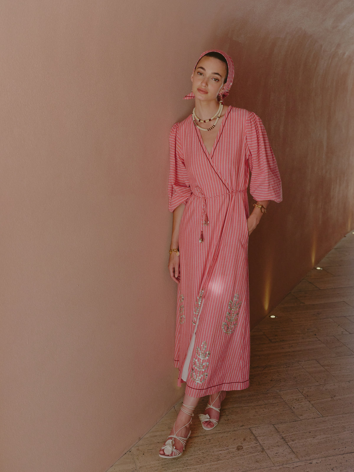 Model wearing a pink and white striped wrap dress with embroidered details and a matching headscarf, standing in a curved hallway.