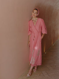 Model wearing a pink and white striped wrap dress with embroidered details and a matching headscarf, standing in a curved hallway.
