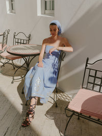 Woman sitting at a small outdoor café table wearing a strapless blue and white striped midi dress with embroidered details and a matching headscarf, styled with black lace-up sandals.