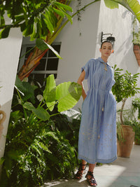 Full-length view of a woman wearing a blue and white striped collared midi dress with embroidered details, styled with black lace-up sandals and a gold anchor necklace, standing among lush greenery in a courtyard.