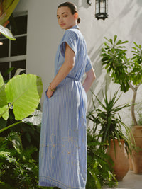 Back view of a woman wearing a blue and white striped midi shirt dress with embroidered patterns near the hem, standing in a sunlit garden with tropical plants.