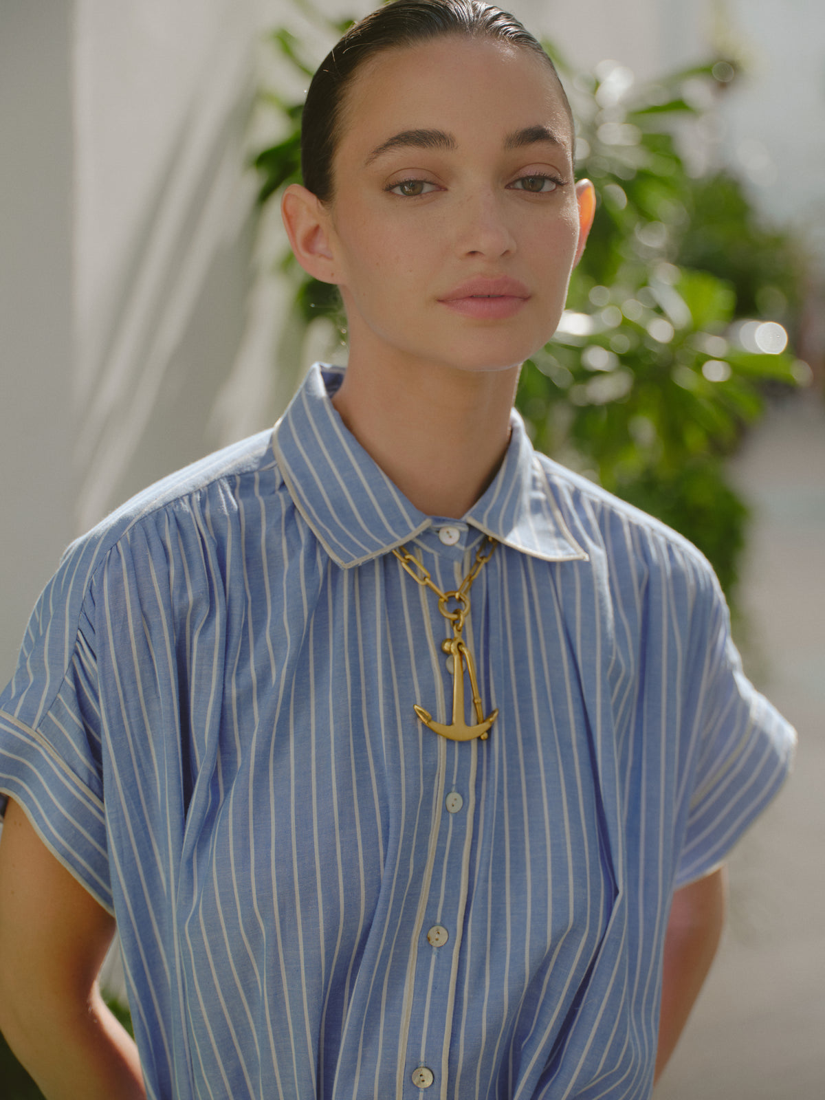Close-up portrait of a woman wearing a blue and white striped collared shirt dress and a chunky gold anchor necklace, with soft sunlight and greenery in the background.