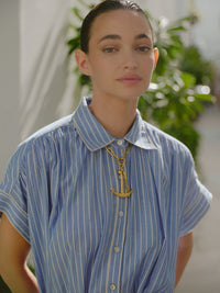 Close-up portrait of a woman wearing a blue and white striped collared shirt dress and a chunky gold anchor necklace, with soft sunlight and greenery in the background.