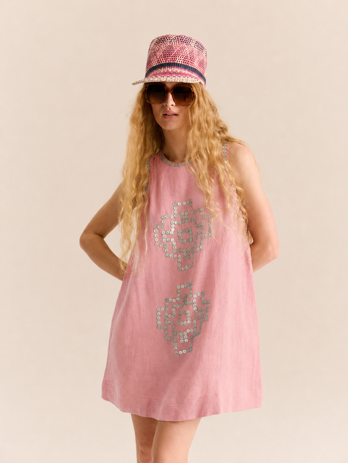 Model wearing a sleeveless pink linen shift dress with a relaxed A-line silhouette. The dress features hand-applied sequin embellishments in geometric motifs down the front and around the neckline and armholes. Styled with oversized brown sunglasses, a pink woven bucket hat with contrasting stripes, and tan woven flats for a playful summer look.