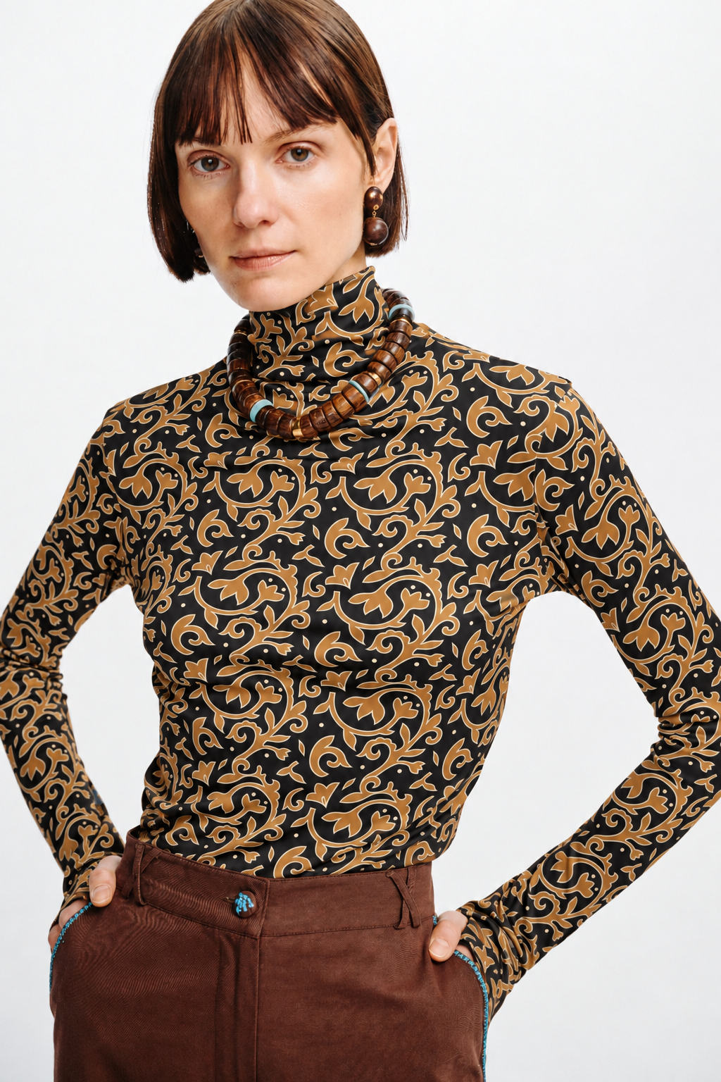 Model wearing a Carolina K charcoal turtleneck with a caramel-brown ornamental floral print, styled with brown pants and wooden jewelry.