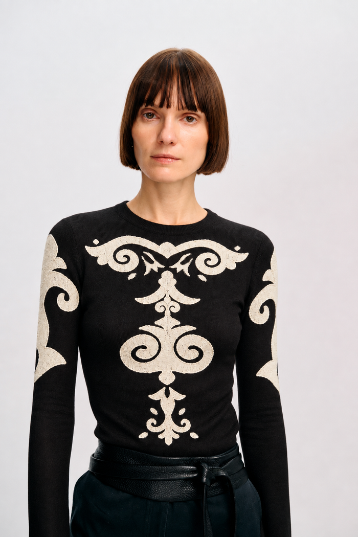 Model wearing the black Elizabeth Top with ivory ornamental appliqué, styled with black trousers and a wrap belt.