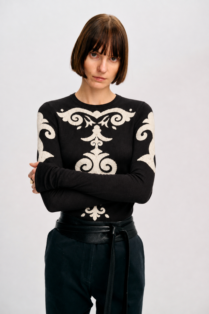 Model posing with crossed arms in the black Elizabeth Top, featuring ivory appliqué motifs across the front and sleeves.