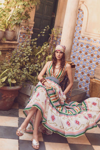 Model seated in a tiled courtyard wearing a colorful striped maxi dress with geometric patterns, layered gold jewelry, and a printed headscarf.