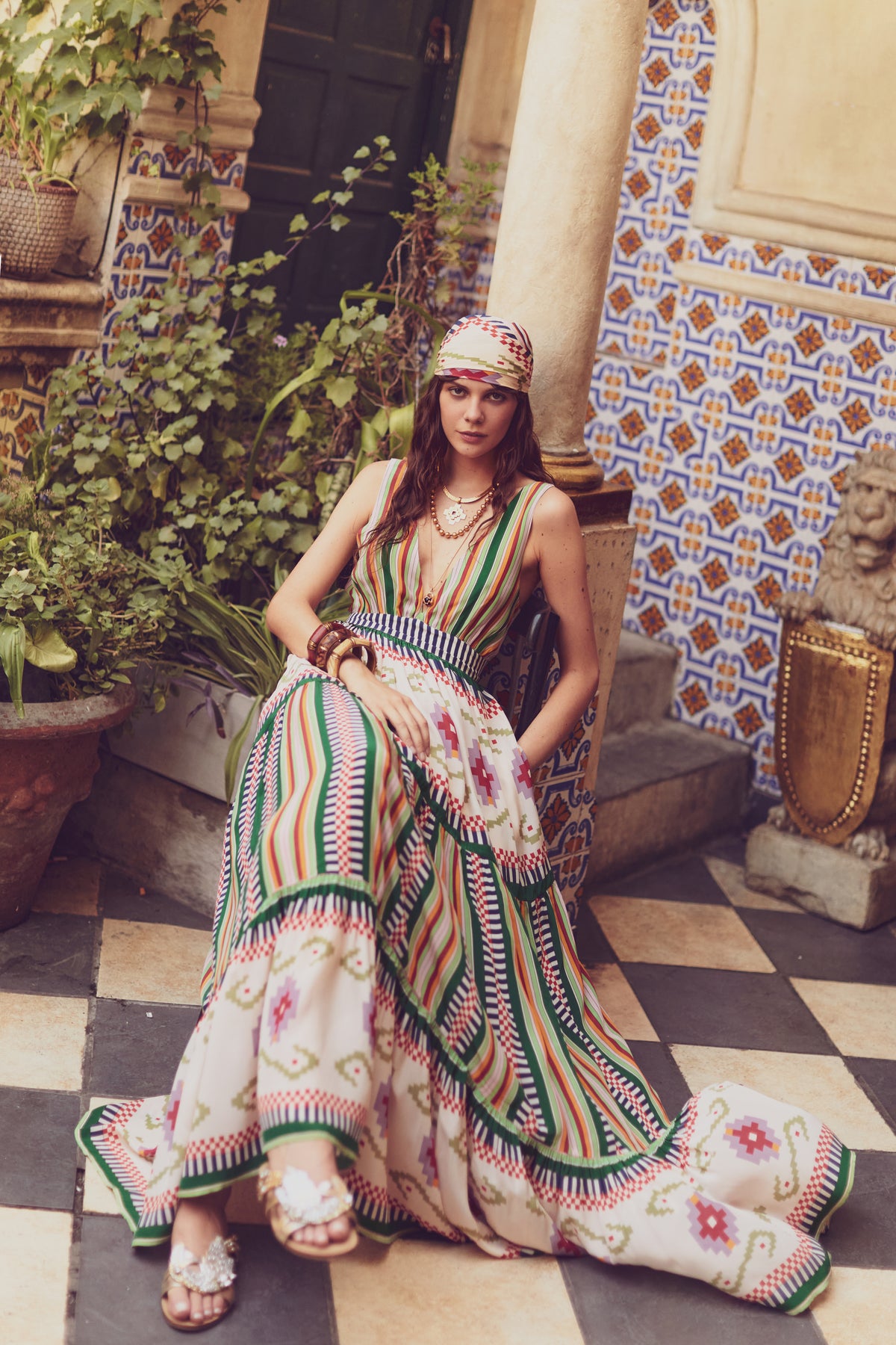 Model sitting in a Spanish-style courtyard wearing a striped and geometric print maxi dress with a deep V neckline, styled with gold jewelry and a headscarf.