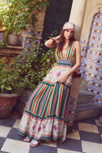 Model standing beside a column in a Mediterranean courtyard wearing a tiered striped maxi dress with geometric motifs and a matching headscarf.