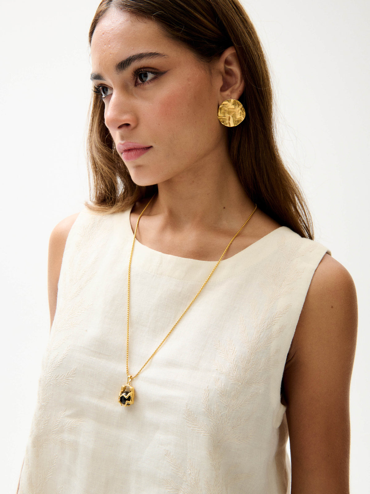 Close-up of cream embroidered sleeveless top with round neckline styled with gold pendant necklace and earrings