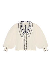 Front view of a cream blouse with a large embroidered collar, navy floral detailing, sheer puff sleeves, and embroidered trim along the button placket and cuffs.