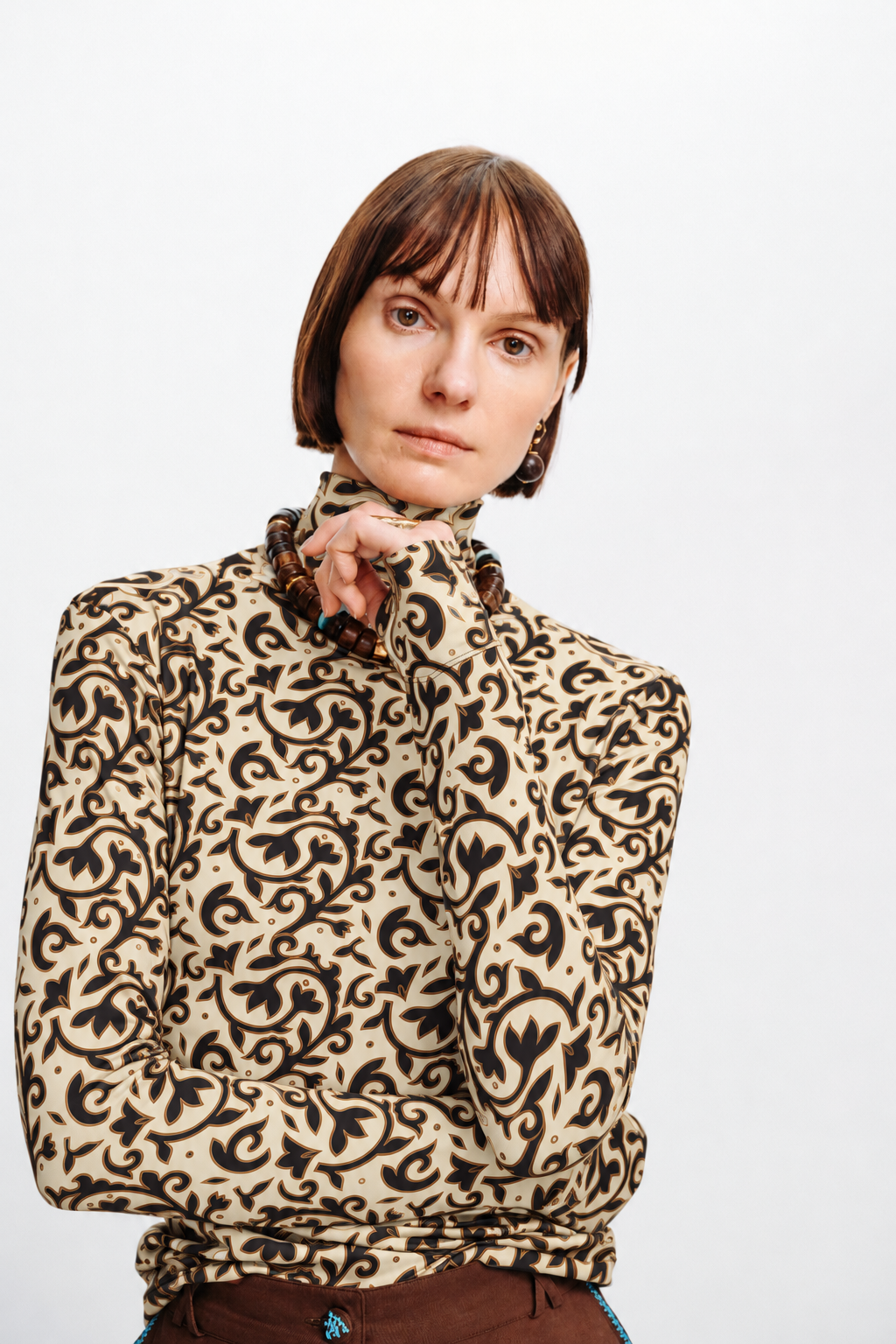 Model wearing an ivory turtleneck with a dark brown floral motif, styled with brown pants and statement jewelry.