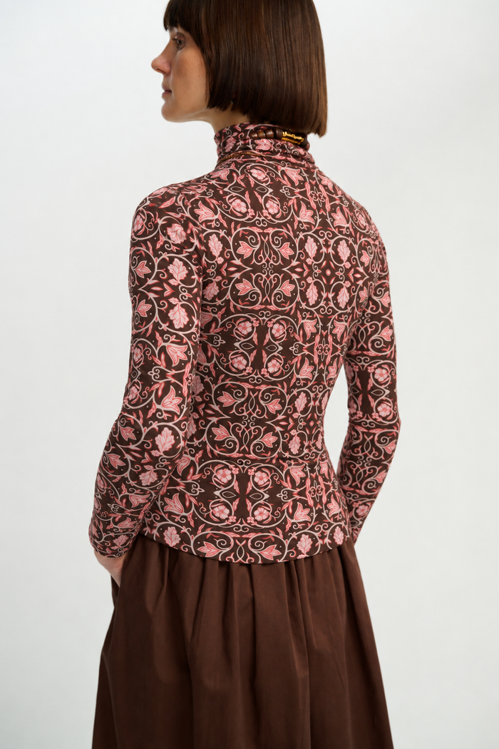 Back view of a model wearing the fitted chocolate-brown turtleneck with an all-over pink botanical print, styled with a brown skirt.
