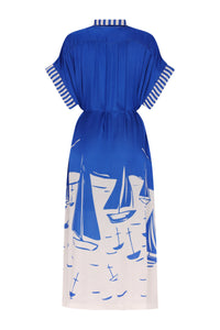 Royal blue midi kaftan dress with a relaxed silhouette, short wide sleeves, and an adjustable drawstring waist. The back features striped trim on the collar and sleeve cuffs, while the lower skirt is adorned with a bold white sailboat print inspired by a coastal marina scene.